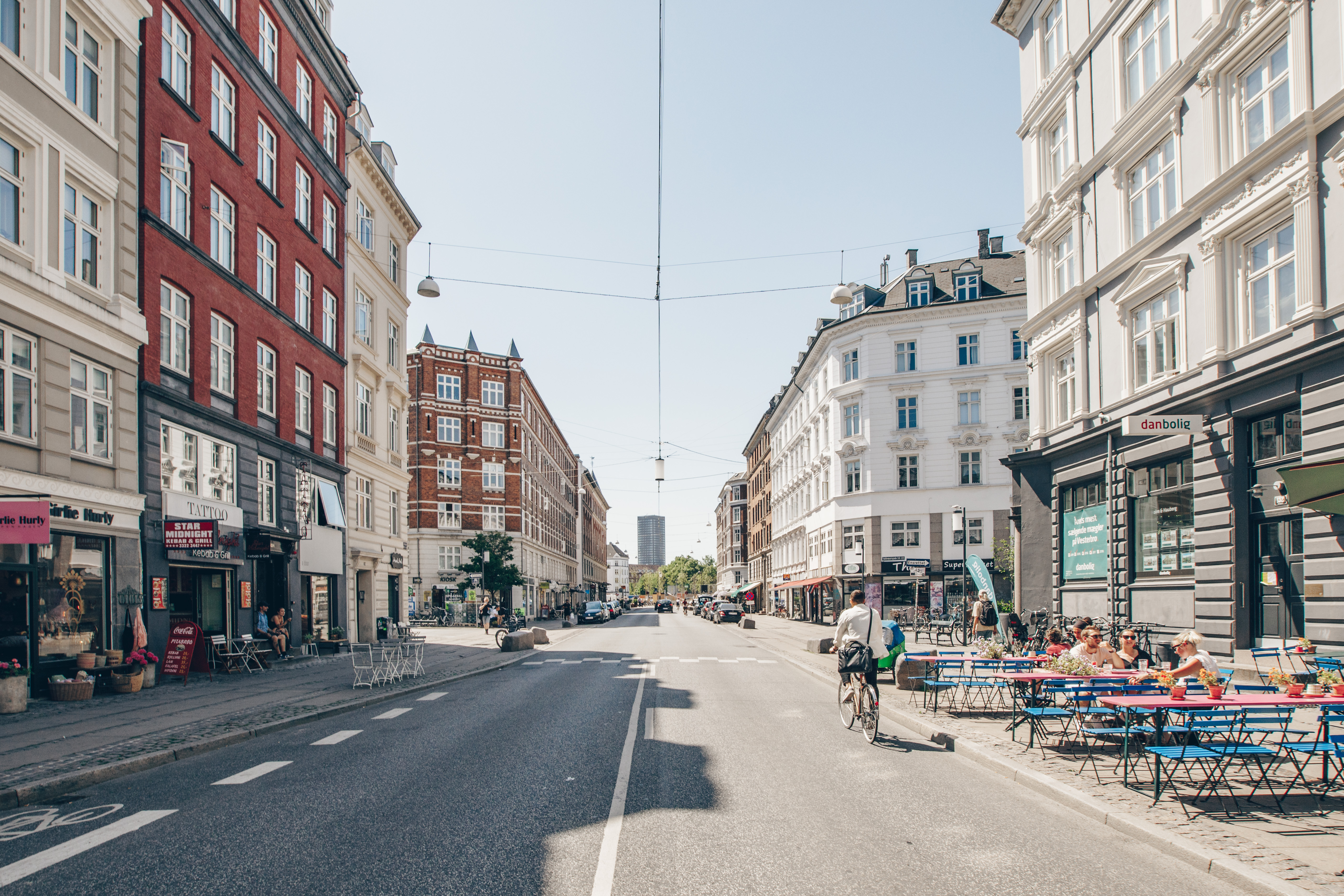 A vibrant street scene featuring colorful buildings, outdoor seating, and cyclists. Sunlight illuminates the lively atmosphere of the area.