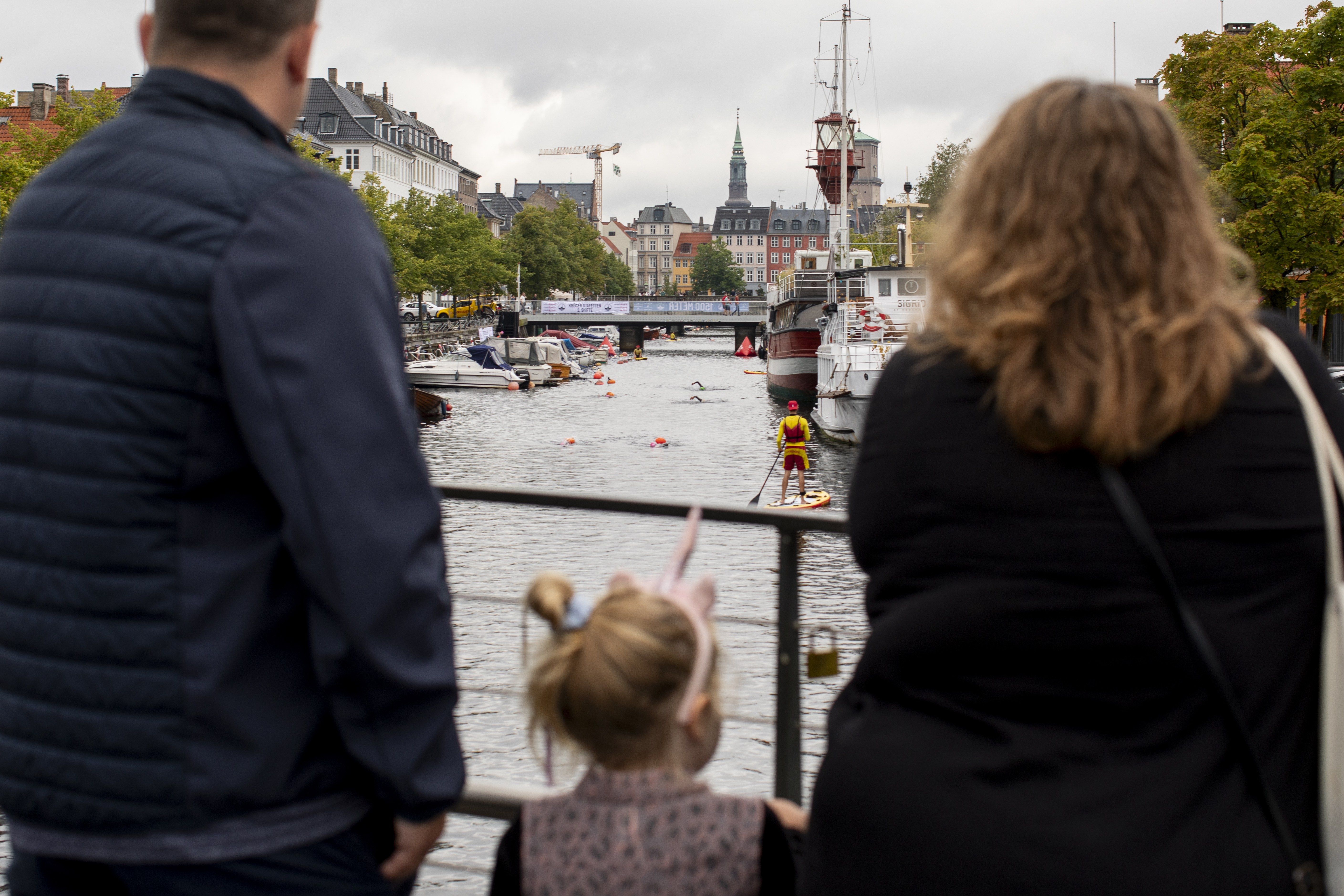 A family watches a canal scene with boats and a paddleboarder. Historic buildings and trees line the waterway, creating a vibrant atmosphere.