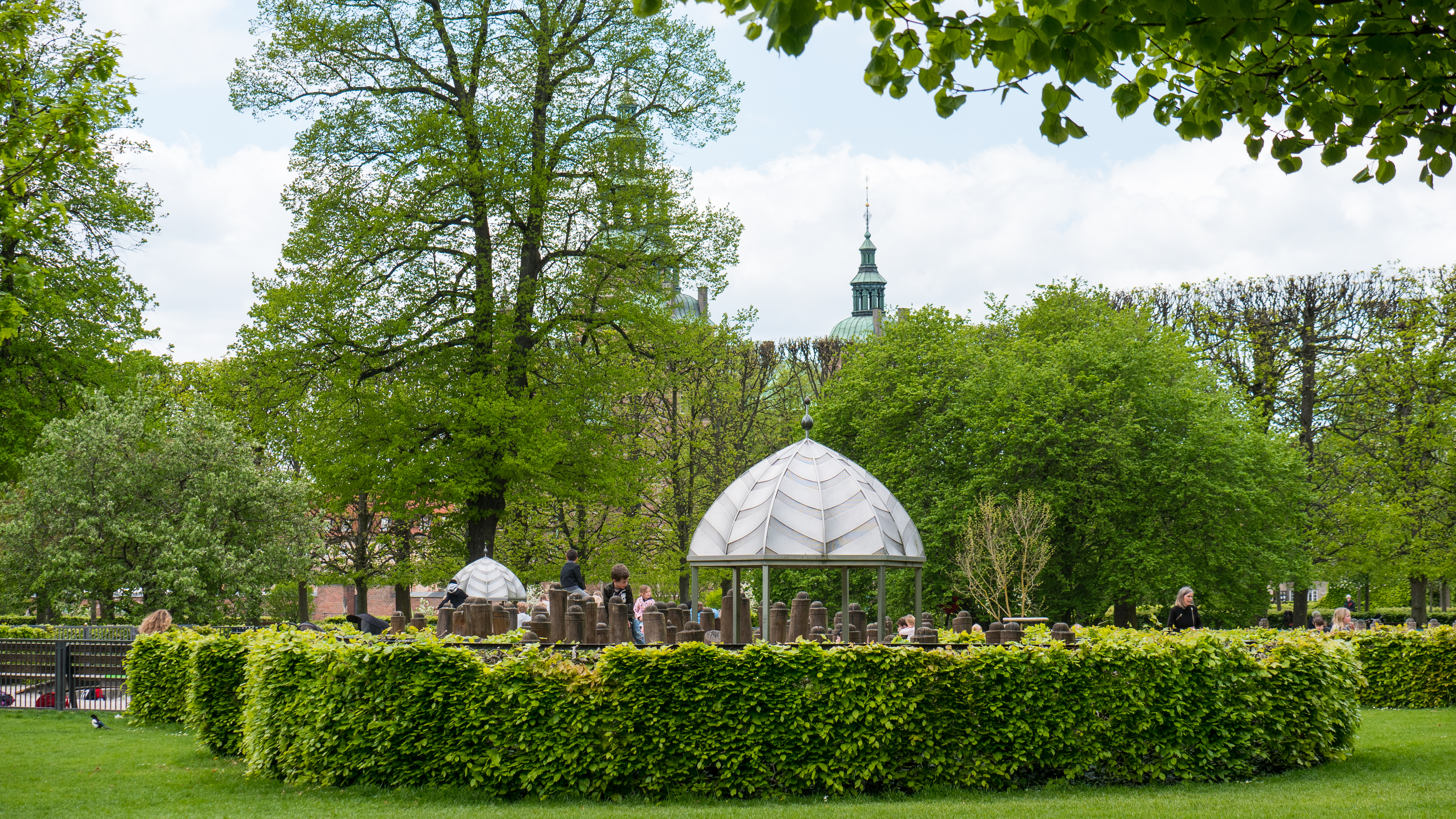 Lush green park featuring a circular hedge surrounding a gazebo, with visitors enjoying the serene landscape and trees in the background.