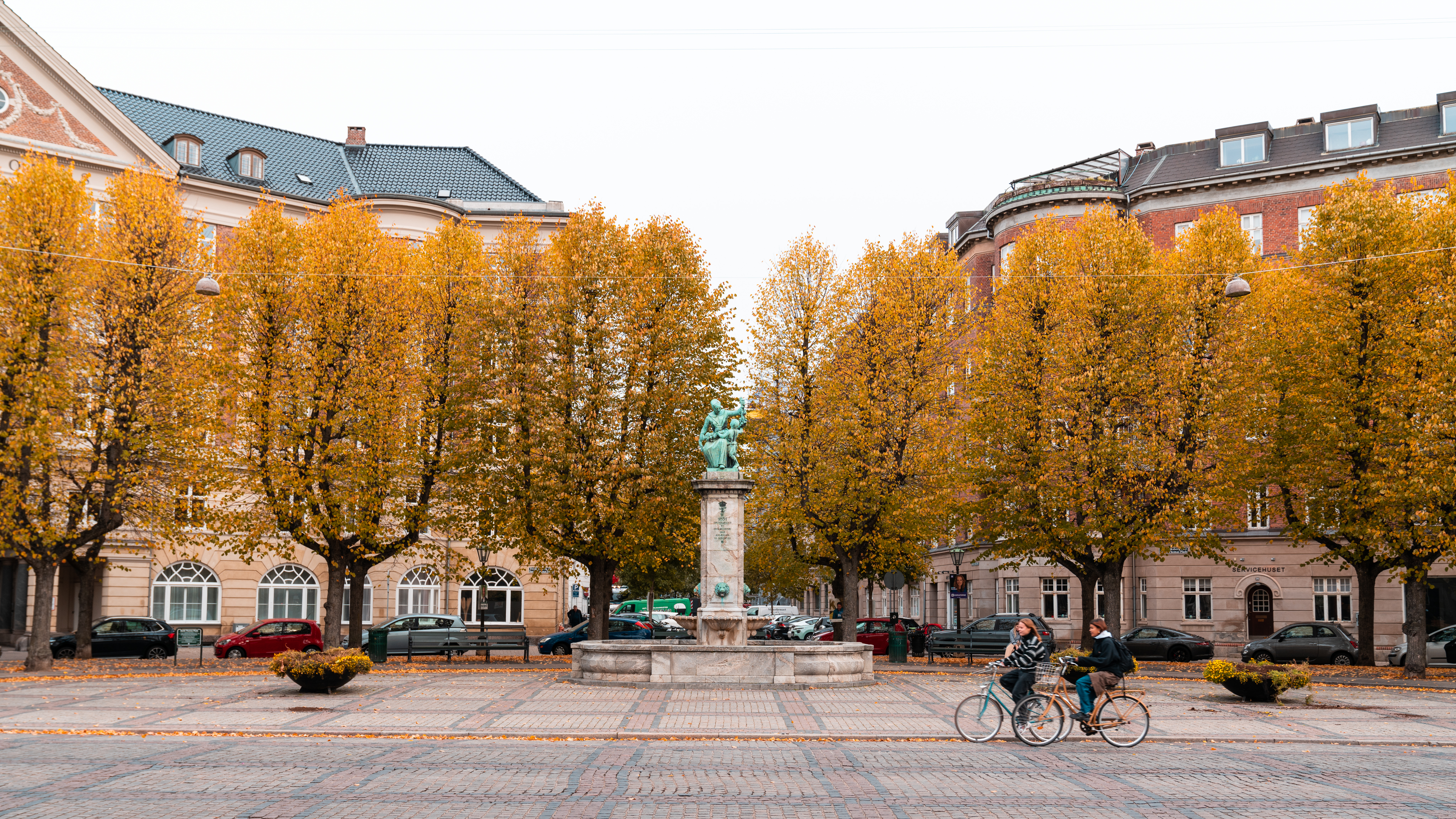 A public square lined with golden autumn trees, featuring a statue at the center. Two cyclists pass by, with historic buildings in the background.