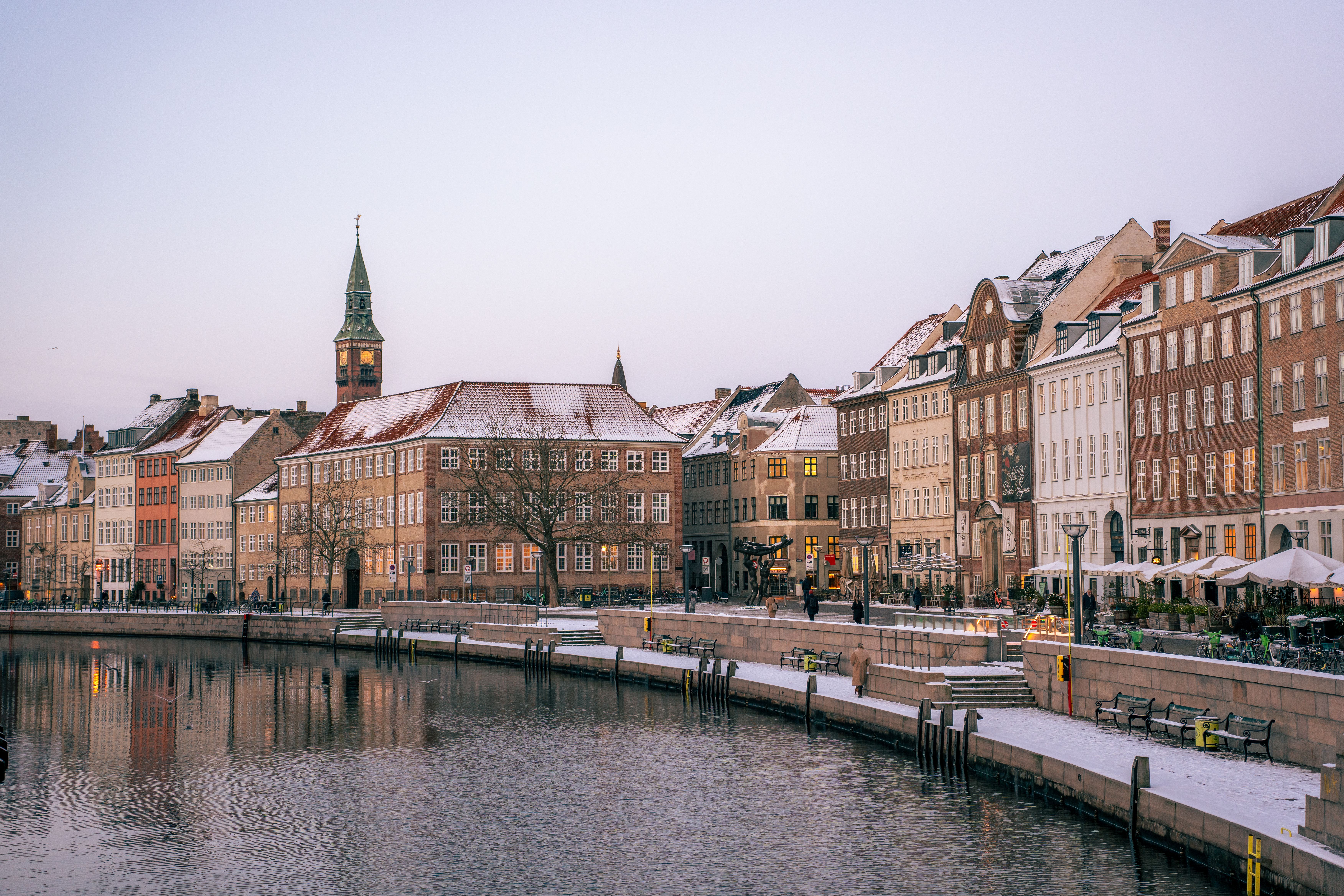 Scenic view of Gammel Stand lined with historic buildings, a clock tower, and snow-dusted rooftops at dusk with lights reflecting in the water.