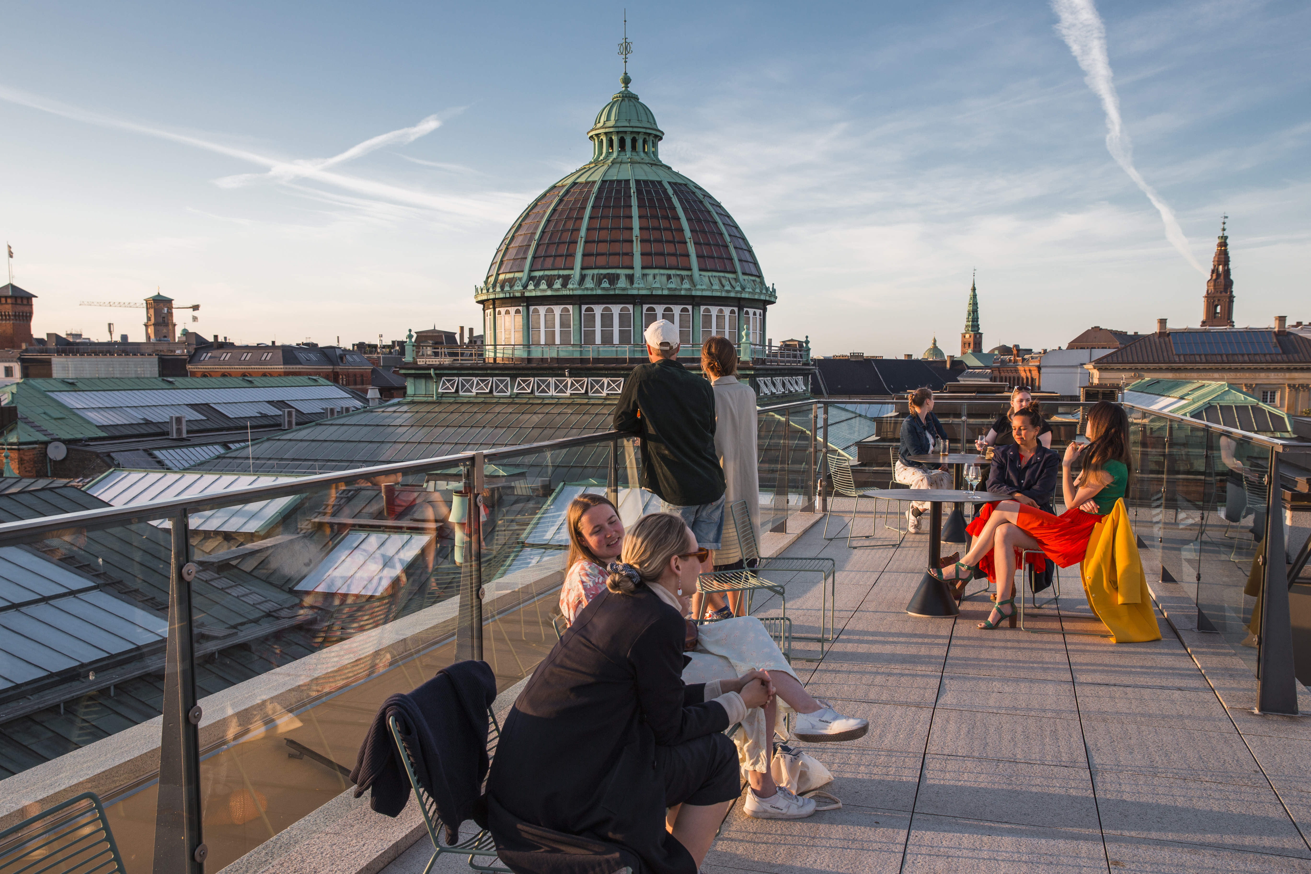 Rooftop terrace with people enjoying the view of a city skyline, featuring a large domed building and vibrant sunset sky.